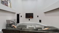 This single-floor space centers around a thoughtfully designed media lounge that prioritizes comfort and entertainment. The room immediately draws attention with its dramatically high ceilings, which lend an expansive, open feel to the interior. Neutral light gray walls create a clean, modern backdrop that complements the space's minimalist aesthetic.

A large wall-mounted flat-screen television serves as the focal point, flanked by built-in in-wall speakers positioned strategically throughout the room to deliver a full surround sound experience. Additional speakers are mounted at varying heights along the walls, reinforcing the space's dedication to high-quality audio performance.

The seating arrangement features a generous sectional sofa in a muted gray fabric, accompanied by a central ottoman that doubles as a coffee table. Accent pillows in warm tones provide a subtle contrast to the otherwise cool palette. Soft carpeting underfoot adds a layer of comfort suited to extended viewing sessions.

A black-framed door on the left side of the room provides access to an adjacent area, while a small seating nook is partially visible beyond it, suggesting additional functional space nearby. Overall, the room balances modern design with practical amenities.