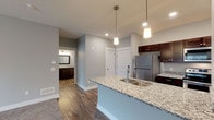 This single-floor residence offers a thoughtfully designed open-concept layout that seamlessly connects the living and kitchen areas. The kitchen is a standout feature, equipped with rich dark-stained cabinetry, granite countertops, stainless steel appliances including a refrigerator, range, and over-the-range microwave, and a central island with a built-in sink and pendant lighting overhead. Wood-look flooring runs throughout the kitchen and main living corridor, adding warmth and durability to the space.

The living area is generously sized, finished with plush carpeting and a ceiling fan, and features a sliding glass door that allows natural light to pour in. The bedrooms continue the carpeted flooring and ceiling fan motif, each offering closet storage and windows with blinds for privacy and light control.

The bathroom is well-equipped with a granite-topped vanity, dark cabinetry, a full tub-shower combination, and the added convenience of a stacked Whirlpool washer and dryer unit. Neutral gray walls throughout the residence create a clean, contemporary backdrop that complements the consistent finishes across every room, resulting in a cohesive and move-in-ready living space.