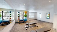 This compact yet fully functional fitness room offers a well-organized workout environment designed to support a variety of exercise routines. The space is finished with durable textured carpet flooring throughout, contributing to a clean and practical aesthetic. Recessed ceiling lighting provides consistent, even illumination across the room, while multiple windows fitted with white horizontal blinds allow natural light to filter in and offer views of surrounding greenery.

A large wall-mounted mirror runs along one side of the room, enhancing the sense of openness and allowing users to monitor their form during workouts. Equipment is thoughtfully arranged throughout the space, including a multi-tiered rack stocked with colorful exercise balls of varying sizes, a dumbbell rack with a full range of free weights, and multiple adjustable weight benches positioned for easy access. A yoga/stretching mat is mounted on the wall alongside a printed guide for stretching poses.

Climate comfort is ensured by a wall-mounted mini-split air conditioning unit, making the space suitable for year-round use. A sleek stainless steel waste bin adds a polished, functional touch. The bright white walls and textured ceiling contribute to the room's clean, modern feel. Altogether, this fitness room is a practical and inviting space for strength training, stretching, and general exercise.