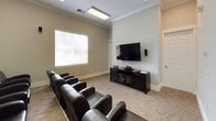 This single-floor media room is thoughtfully designed as a private screening and entertainment space. The room is arranged with multiple rows of dark leather club chairs positioned to face a large wall-mounted flat-screen television, supported by a dark wood media console housing audio-visual equipment. The seating arrangement accommodates a generous number of viewers, making it well-suited for group screenings or gatherings.

The room features neutral-toned walls accented by crisp white crown molding and white baseboards, lending a clean, polished aesthetic. Soft, neutral-toned carpeting runs throughout, contributing to the room's acoustically suitable environment. Overhead, recessed and flush-mounted light fixtures provide ample illumination, while window blinds along the exterior-facing walls allow for flexible light control during viewings.

A notable decorative feature is a curated gallery wall of black-and-white photographic prints depicting iconic pop culture figures, adding a stylish, cinematic character to the space. Wall-mounted speakers are positioned strategically to support an immersive audio experience. Overall, the space combines functional media room design with a comfortable, lounge-like ambiance ideal for entertainment.