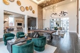 This thoughtfully designed single-floor communal space blends contemporary elegance with functional social areas across several distinct zones. The central lounge commands immediate attention with its dramatic double-height tray ceiling, large-scale sputnik chandelier, and floor-to-ceiling windows that flood the interior with natural light while offering outdoor views. Comfortable seating — including a white sofa, tufted leather chairs, and a geometric faceted coffee table — anchors this gathering area, with a billiards table visible in the background.

Adjacent to the main lounge, a café-style bar area features an elongated waterfall-edge marble island with wood-and-cane barstools, bold geometric gold pendant lights, and a textured dark herringbone accent wall behind sleek gray cabinetry with marble countertops and stainless steel appliances, including a Starbucks coffee station.

A dedicated dining area showcases a live-edge wood table surrounded by velvet chairs beneath a globe chandelier, complemented by a striking vertical patterned accent wall and floor-to-ceiling windows overlooking an outdoor courtyard.

A cozy lounge nook features built-in charcoal velvet banquette seating with orange accent pillows, paired with swivel chairs and marble-top side tables atop a geometric-patterned rug, with curated gallery walls throughout adding an artistic, refined atmosphere.