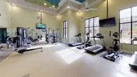 This fitness center is a thoughtfully designed workout space that accommodates a comprehensive range of exercise options. The room features soaring ceilings, which contribute to an open and airy atmosphere, complemented by multiple ceiling fans that promote air circulation throughout. The walls are painted in a soft sage green, creating a calm and motivating environment.

Large, black-framed windows line the walls, flooding the space with natural light and offering views of the surrounding outdoor courtyard and building complex. The durable, light-toned flooring provides a clean and functional surface throughout.

The cardio section is well-stocked with multiple treadmills, an elliptical machine, a stationary recumbent bike, and a rowing machine. A dedicated strength-training area includes a multi-station cable machine, a weight bench, and a tiered dumbbell rack with a full range of free weights. Full-length mirrors along one wall visually expand the space while allowing users to monitor their form during workouts.

Multiple wall-mounted flat-screen televisions are positioned throughout for entertainment during exercise. Recessed lighting and a clearly marked exit door complete this practical, well-appointed fitness facility.