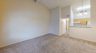 This single-story space is characterized by a practical, open layout spread across two distinct zones visible in the floor plan. The living area makes a strong impression with its soaring vaulted ceilings, which lend a sense of spaciousness beyond the room's physical footprint. A tile-surround fireplace serves as the focal point of this room, complemented by a closet and a front entry door. A window alongside the entry admits natural light and offers a view of surrounding greenery.

Carpeting in a neutral, medium-toned beige runs throughout the living and bedroom areas, providing a cohesive feel across the space. The bedrooms feature textured ceilings and windows that frame outdoor tree views, enhancing the connection to the exterior environment. Each carpeted bedroom provides a calm, neutral backdrop suitable for a range of furnishing styles.

A hallway connects the living zone to the private areas of the home, leading past a kitchen space with wood-toned cabinetry visible from the 3D floor plan. The neutral wall palette throughout — soft whites and warm beige tones — creates a clean, understated ambiance. HVAC venting is integrated into the walls, and the overall layout prioritizes straightforward, functional living on a single level.