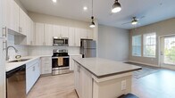 Located at 2035 Tapestry Park Drive in Indian Land, SC, this well-appointed one-bedroom, one-bathroom residence offers a thoughtfully designed open-concept layout with modern finishes throughout. The combined kitchen and living area serves as the heart of the home, featuring light wood-style flooring, soaring ceilings, and abundant natural light streaming in through large windows and a glass-panel exterior door. The kitchen is outfitted with crisp white shaker-style cabinetry, a classic subway tile backsplash, quartz countertops, a central island with pendant lighting, and a full suite of stainless steel appliances including a range, microwave, dishwasher, and refrigerator. The living area provides an open and airy feel with high ceilings and direct outdoor access. The bedroom is generously sized with plush carpeting, elevated ceilings, and windows that bring in natural light. It connects to a walk-in closet with built-in wire shelving for ample storage. The bathroom features light wood-tone cabinetry, a wide quartz-topped vanity, a large mirror, and wood-look flooring, creating a clean and contemporary feel throughout. The neutral color palette and modern design elements give the entire space a cohesive, move-in-ready aesthetic.