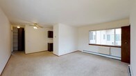 This single-floor residential space offers a clean and functional layout designed for comfortable everyday living. The main living area is finished with wall-to-wall carpet flooring, providing a soft and warm underfoot feel throughout. A ceiling fan is centrally mounted overhead, ensuring air circulation across the space. A large sliding window allows natural light to fill the room, creating a bright and welcoming atmosphere, while a baseboard heating unit beneath it ensures year-round comfort. The neutral white walls keep the space feeling open and versatile, ready to accommodate a variety of furniture arrangements and décor styles. A partial dividing wall separates the living area from the kitchen, offering a degree of definition between the two spaces while maintaining an open feel. The kitchen features dark wood cabinetry, providing a warm contrast to the lighter tones of the living area. The overall layout is straightforward and practical, making it easy to navigate and utilize. The space presents a functional, no-frills residential environment that is well-suited for those seeking a simple, move-in-ready home.