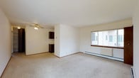This single-floor residential space offers a clean and functional layout designed for comfortable everyday living. The main living area is finished with wall-to-wall carpet flooring, providing a soft and warm underfoot feel throughout. A ceiling fan is centrally mounted overhead, ensuring air circulation across the space. A large sliding window allows natural light to fill the room, creating a bright and welcoming atmosphere, while a baseboard heating unit beneath it ensures year-round comfort. The neutral white walls keep the space feeling open and versatile, ready to accommodate a variety of furniture arrangements and décor styles. A partial dividing wall separates the living area from the kitchen, offering a degree of definition between the two spaces while maintaining an open feel. The kitchen features dark wood cabinetry, providing a warm contrast to the lighter tones of the living area. The overall layout is straightforward and practical, making it easy to navigate and utilize. The space presents a functional, no-frills residential environment that is well-suited for those seeking a simple, move-in-ready home.