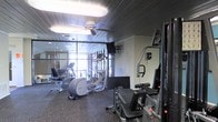This fitness center is designed to support a well-rounded workout routine, offering a thoughtfully arranged layout with a diverse selection of exercise equipment. The space features several cardio machines, including an elliptical trainer and a stair climber, alongside strength training equipment such as a cable machine and weight bench, providing users with comprehensive training options. Rubber flooring throughout the space offers both safety and durability, making it well-suited for high-intensity activities. Mirrored walls line one side of the room, visually expanding the space while allowing users to monitor their form during workouts. Large windows along the front of the facility invite natural light and provide an open, airy feel. A ceiling fan overhead contributes to air circulation and overall comfort during exercise. The paneled ceiling adds a distinct visual character to the space. Wall-mounted screens provide entertainment or instructional content during workouts. The overall ambiance is functional and practical, with a clean, organized arrangement of equipment that makes efficient use of the available floor space, catering to a variety of fitness needs and experience levels.