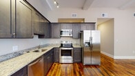 Located at 89 Globe Mills Ave in Fall River, MA, this single-floor residence blends historic mill building character with modern finishes throughout. Rich, original hardwood floors run continuously across the open living and kitchen areas, lending warmth and a sense of industrial heritage to the space.

The kitchen is well-appointed with dark-stained shaker-style cabinetry, granite countertops, and a full suite of stainless steel appliances including a range, over-the-range microwave, dishwasher, and a side-by-side refrigerator with ice and water dispenser. Track lighting illuminates the workspace, while the layout maximizes storage and counter space efficiently.

The open living area is a standout, featuring soaring ceilings, exposed structural beams, a ceiling fan, and a dramatic wall of oversized arched windows that flood the room with natural light and provide an impressive architectural focal point. The generous proportions of this area offer significant flexibility for living and dining arrangements.

The bedroom includes hardwood flooring, a closet with built-in shelving, and direct access to additional rooms. The full bathroom is finished with neutral tile flooring, a dark wood vanity with granite countertop, a large mirror, and a shower-tub combination. An in-unit stacked washer and dryer adds everyday convenience to this well-designed space.