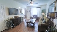 This single-floor home offers a warm and inviting atmosphere anchored by rich hardwood flooring that flows throughout the main living area. The living room is generously proportioned, featuring a ceiling fan with integrated lighting that enhances both comfort and functionality. Multiple windows dressed with elegant draping curtains allow abundant natural light to fill the space, creating a bright and airy ambiance. A large flat-screen television is mounted on the wall above a dark, ornately carved storage credenza, which serves as a striking focal point. The space is thoughtfully furnished with a classic upholstered sofa, a sturdy wooden coffee table with turned legs, and a cozy accent chair positioned near the windows. Decorative touches such as potted greenery, tall bamboo plants, and carefully placed candle holders add character and warmth to the room. The neutral-toned walls and coordinated décor contribute to a cohesive, relaxed aesthetic. Overall, the home presents a comfortable and well-appointed living environment that balances traditional design elements with everyday practicality.