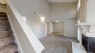This two-story space is defined by its dramatic vaulted ceilings, which lend an expansive, airy quality to the main living area. The living room serves as the focal point of the lower level, showcasing a striking floor-to-ceiling shiplap-accented fireplace with a tiled hearth and wood mantle. Hardwood-style luxury vinyl flooring runs throughout the main living and dining areas, complementing the neutral warm-beige walls. A staircase with white railings leads to the upper level, where the soaring ceiling continues to impress.

The kitchen is well-appointed with white cabinetry, quartz countertops, a subway tile backsplash, and stainless steel appliances including a range and built-in microwave. A skylight adds natural brightness to the space. Adjacent areas provide additional storage, including a dedicated pantry with built-in shelving.

The bedroom features plush carpeting, a ceiling fan, and a window with blinds that allows natural light. The bathroom includes a full tub-shower combination, toilet, and overhead cabinetry for storage, finished with matching hardwood-style flooring. A vanity area with quartz countertops and access to a linen closet adds practical functionality.