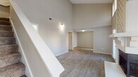 This two-story space is defined by its dramatic vaulted ceilings, which lend an expansive, airy quality to the main living area. The living room serves as the focal point of the lower level, showcasing a striking floor-to-ceiling shiplap-accented fireplace with a tiled hearth and wood mantle. Hardwood-style luxury vinyl flooring runs throughout the main living and dining areas, complementing the neutral warm-beige walls. A staircase with white railings leads to the upper level, where the soaring ceiling continues to impress.

The kitchen is well-appointed with white cabinetry, quartz countertops, a subway tile backsplash, and stainless steel appliances including a range and built-in microwave. A skylight adds natural brightness to the space. Adjacent areas provide additional storage, including a dedicated pantry with built-in shelving.

The bedroom features plush carpeting, a ceiling fan, and a window with blinds that allows natural light. The bathroom includes a full tub-shower combination, toilet, and overhead cabinetry for storage, finished with matching hardwood-style flooring. A vanity area with quartz countertops and access to a linen closet adds practical functionality.