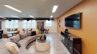 This single-floor communal space is thoughtfully designed to serve as a gathering and entertainment hub. The lounge area immediately captures attention with its bold amber accent wall, which provides a warm, energetic backdrop to the carefully curated décor, including woven wall art and decorative platters. A large wall-mounted flat-screen television anchors the entertainment setup, complemented by a curved sectional sofa, a round textured coffee table, and additional seating options that encourage relaxed social interaction. Recessed rectangular panel lighting illuminates the space evenly, while light wood-look flooring runs seamlessly throughout, tying the areas together. Large windows along one wall invite natural light and offer views of the surrounding landscape, enhancing the open and airy ambiance.

The adjoining kitchen is equally impressive, featuring rich dark wood cabinetry with sleek bar-pull hardware, granite-style countertops, and a classic white subway tile backsplash. A stainless steel refrigerator and a double-basin sink add functional utility, while a substantial central island with pendant lighting — highlighted by two striking geometric cage-style fixtures — provides ample prep and serving space. The overall design balances contemporary style with practical amenities, making this space well-suited for community events, gatherings, and everyday socializing.