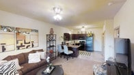 This single-floor residence is designed with an open-concept layout that creates a cohesive and inviting atmosphere throughout. The living area features warm, neutral-toned carpet flooring and is centered around a cozy seating arrangement, complemented by a decorative shelving unit and tasteful wall art that add character to the space. Ceiling fans with light fixtures provide both illumination and comfort throughout the unit.

The kitchen is a standout feature, showcasing rich dark-stained cabinetry that extends to the upper walls, offering generous storage space. Black appliances, including a refrigerator, stove, and microwave, lend a sleek, modern look to the culinary space. A kitchen island with bar seating for two serves as a natural divider between the kitchen and living area, making it ideal for casual dining or entertaining.

The transition between spaces is smooth and natural, with the open sightlines creating a sense of spaciousness. The textured ceiling and soft, warm lighting throughout contribute to an overall ambiance that is both functional and welcoming. The layout is thoughtfully organized to maximize usability while maintaining an open and airy feel.