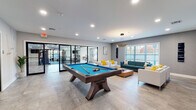 Located at 55 W Merritt Blvd in Fishkill, NY, this single-floor amenity center offers a thoughtfully designed space that combines social and fitness-oriented areas in a modern, cohesive layout. The lounge area serves as a welcoming gathering space, featuring a teal sofa, white armchairs accented with yellow throw pillows, and a warm wood coffee table arranged beneath a striking circular gold pendant light fixture. A wall-mounted flat-screen TV and textured gray accent walls complete the polished aesthetic, while large windows invite abundant natural light and views of the surrounding landscape. Adjacent to the lounge, a billiards area centers around a full-size pool table with a teal felt surface, positioned atop large-format stone-look tile flooring that extends throughout the space. The fitness center is accessible through sleek glass and metal doors, and is divided into distinct zones. A cardio and functional training area features treadmills, a stationary bike, resistance bands, and yoga mats, along with a wall-mounted TV and mirrored walls to enhance the sense of space. A separate strength training zone houses a comprehensive selection of cable machines, free weights, and benches, all under recessed lighting with ample room to move freely. Clean, modern finishes, light wood flooring in the gym areas, and generous window placement throughout create a bright, energizing environment ideal for both activity and relaxation.