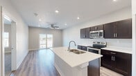 This well-designed single-floor unit showcases a clean, contemporary aesthetic throughout. The open-concept layout seamlessly connects the kitchen and living area, creating a bright and functional shared space. The kitchen is a standout feature, equipped with rich dark cabinetry, white quartz countertops, and a full suite of stainless steel appliances including a range, built-in microwave, and a side-by-side refrigerator with an ice and water dispenser. A generously sized center island with a double-basin sink serves as both a prep area and a natural gathering point. Light wood-look flooring runs throughout the main living areas, complementing the neutral gray walls and recessed lighting. The living area benefits from abundant natural light and is anchored by a ceiling fan, with French doors opening onto a private balcony offering open outdoor views. The bedrooms feature plush carpet flooring and are finished in soft, neutral tones, providing a calm and comfortable retreat. The bathroom is outfitted with dark wood vanity cabinetry consistent with the kitchen's design, wood-look tile flooring, and clean white fixtures. A laundry area is also present within the unit. Overall, the space presents a cohesive, modern finish with practical, well-appointed details throughout.