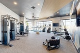 Located in Aurora, IL, this well-appointed fitness center offers a generously sized, single-floor exercise space designed to accommodate a full spectrum of workout routines. The facility features a contemporary aesthetic with a striking combination of dark ceiling panels, warm wood slatted accents, and recessed lighting complemented by ceiling fans throughout.

The cardio zone is equipped with Matrix treadmills, elliptical trainers, stair climbers, and stationary bikes, all positioned facing large, floor-to-ceiling windows that frame open outdoor views, creating an energizing and bright atmosphere for exercise. A wall-mounted flat-screen TV adds to the amenity.

The strength training area includes a cable crossover machine, a squat rack with weight plates, selectorized machines, dumbbells, medicine balls, and stability balls neatly organized along a full-length mirror wall. A heavy punching bag and functional training tools such as a battle rope and large tire highlight the facility's dedication to diverse training modalities.

A dedicated barre studio section features ballet barres set against floor-to-ceiling mirrors and hardwood flooring, ideal for group fitness or individual training. A bold abstract mural serves as a dynamic visual centerpiece. The overall ambiance is modern, motivating, and professionally outfitted.