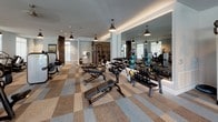 This modern fitness center is thoughtfully designed to accommodate a wide range of workout styles and fitness goals. The main exercise floor is finished with a distinctive checkered carpet tile pattern and is generously illuminated by pendant lighting, creating a focused yet inviting atmosphere. Large windows along the perimeter flood the space with natural light and offer views of the surrounding outdoors.

The cardio zone is equipped with stationary bikes, cycling bikes with interactive screens, and a stair climber machine, all positioned to take advantage of the natural light from the expansive windows. Nearby, a comprehensive free weights area features multiple racks of dumbbells, kettlebells, medicine balls in assorted weights, and adjustable weight benches, all reflected in floor-to-ceiling mirrors that enhance the sense of space.

A separate functional training area houses a cable machine, stability balls, and step platforms, accessible through glass-panel doors. The studio space is a standout feature, boasting rich hardwood flooring, a ceiling fan, a wall-mounted television, and oversized windows and glass doors that open to outdoor views. Plyo boxes and a stretching area complete this versatile room, making it ideal for group classes, yoga, or open training sessions.