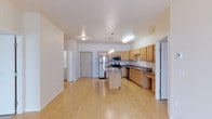 This single-floor space is defined by an open-concept layout that seamlessly connects the living area and kitchen, creating a versatile and functional environment. Light hardwood flooring runs throughout, contributing to a warm, airy atmosphere that reflects natural and artificial light alike.

The kitchen is a standout feature, outfitted with light maple cabinetry that provides generous storage space. Stainless steel appliances — including a refrigerator, range, and built-in microwave — lend a modern, contemporary feel. A central island with a granite-style countertop offers additional workspace and serves as a natural gathering point. The main countertops also feature stone-style surfaces, complementing the overall aesthetic. A pendant light cluster above the island adds focused, stylish illumination, while flush-mount ceiling fixtures ensure the space is well-lit throughout.

The open living area is spacious and unobstructed, offering flexibility for a variety of furniture arrangements. Neutral-toned walls and clean lines maintain a minimalist, welcoming ambiance. A utility area with additional appliances is visible through a doorway, adding to the practical functionality of the unit. The layout and finishes together create a cohesive, move-in-ready space suited to everyday living.