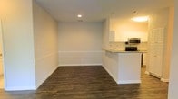 This single-floor residence offers a clean, open-concept layout designed for both comfort and functionality. The living and dining area is finished with rich, dark hardwood-style flooring that contrasts beautifully against the neutral light gray walls, creating a warm yet contemporary ambiance. A classic white chair rail runs along the perimeter of the space, adding a refined architectural detail to the otherwise minimalist design. Recessed lighting in the ceiling provides bright, even illumination throughout the room. The space flows naturally into a modern kitchen, which is separated by a white breakfast bar countertop — ideal for casual dining or entertaining. The kitchen is equipped with white upper cabinetry, a stainless steel microwave, and a black range, all set against a clean white tile backsplash. The countertops offer ample workspace, and the overall palette of white and neutral tones keeps the kitchen feeling bright and airy. The open sightlines between the living, dining, and kitchen areas contribute to a sense of spaciousness, making the layout feel connected and easy to navigate. The consistent flooring, neutral color scheme, and thoughtful architectural accents give this residence a polished, move-in-ready feel.