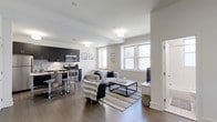 Located along City Avenue in Philadelphia's 19151 zip code, this single-floor residence offers a thoughtfully designed open-concept layout that blends everyday comfort with modern style. The living and kitchen areas flow together seamlessly, creating a spacious and functional environment ideal for both relaxing and entertaining. Rich dark hardwood flooring extends throughout, providing a striking contrast to the bright white walls and light-toned ceilings that keep the space feeling airy and inviting. Natural light pours in through multiple double-hung windows, enhancing the open atmosphere. The kitchen is appointed with dark, sleek cabinetry, granite-style countertops, stainless steel appliances including a range and microwave, and a breakfast bar with modern bar seating — combining practicality with a clean, contemporary aesthetic. The adjacent living area is generously sized, offering ample room for comfortable furnishings arranged around a central coffee table. Recessed and flush-mount ceiling lighting ensures the space remains well-lit throughout the day and evening. A doorway near the living area leads to additional private spaces within the unit. Overall, the residence presents a cohesive, modern design palette with high-quality finishes throughout.