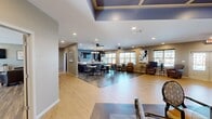 Located at 720 Curtis Pkwy SE in Calhoun, Georgia, this expansive single-floor community space offers a well-designed, open layout that seamlessly connects multiple functional areas. The interior is finished with a mix of light hardwood and dark wood-look flooring, complemented by warm neutral wall tones and recessed lighting throughout.

The centerpiece of the space is a generously sized kitchen featuring bold navy cabinetry, light granite countertops, a large island with pendant lighting, and ample workspace. Ceiling fans and large windows line the kitchen and dining area, flooding the space with natural light. Multiple dining tables provide seating for a sizeable group, making it well-suited for communal gatherings.

Adjacent to the kitchen, the main dining and living area flows openly, with a tray ceiling detail accented in deep blue adding architectural interest overhead. A secondary dining room with double French doors connects to an additional living area, offering flexibility for private events or smaller gatherings. A dedicated lounge area features comfortable leather seating, wall art, and a shuffleboard table, providing a relaxed recreational setting.

Additional living spaces include a bright sitting room with French doors opening to an outdoor patio, as well as a quiet lounge nook with corner windows. The welcoming entry area features clean white panel doors, recessed lighting, and a distinctive blue accent ceiling, setting a polished and inviting tone throughout.