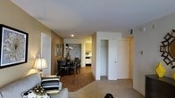 This single-floor residence offers a thoughtfully arranged layout that balances comfort and functionality. The living area is finished with plush carpeting and features recessed lighting in the ceiling, creating a warm and inviting atmosphere. Neutral-toned walls provide a versatile backdrop throughout the space, complemented by warm wood-look flooring in the dining and transition areas. The dining area accommodates a round table with seating for multiple guests and flows naturally from the living space, making it well-suited for everyday meals and entertaining. A glimpse into the kitchen reveals dark cabinetry and countertops with ample workspace, providing a practical and efficient cooking environment. The space includes what appears to be a closet or storage area near the main living zone, adding to the overall utility of the floor plan. Recessed ceiling lights and a table lamp in the living area contribute to a layered lighting scheme. The overall ambiance is cozy and residential, with a clean, move-in-ready aesthetic that suits a variety of lifestyles. The open sightlines between the living, dining, and kitchen zones give the space an airy, connected feel despite its compact footprint.