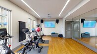 Located at 100 Colonial Homes Dr in Atlanta, GA, this dedicated fitness studio offers a thoughtfully designed workout environment within a compact, single-room layout. The space features warm bamboo hardwood flooring throughout, providing both durability and an inviting aesthetic that contrasts with the clean, neutral-toned walls and ceiling.

Overhead, sleek linear LED lighting runs the length of the ceiling, delivering bright, even illumination across the entire studio. Large windows along one wall allow natural light to filter in, offering views of the surrounding greenery and creating an open, energizing atmosphere.

The room is well-appointed with multiple Peloton spin bikes positioned on protective floor mats, a freestanding ball rack stocked with medicine and stability balls of various sizes, step platforms, a BOSU balance trainer, and free weights. A large wall-mounted flat-screen television anchors one wall, flanked by two mounted speakers that provide an immersive audio experience during workouts.

A full-length mirror spans one wall, visually expanding the space and allowing users to monitor their form. The studio is accessed through louvered double doors as well as a glass-panel entry door equipped with a keypad access system, ensuring secure and convenient entry. The overall ambiance is modern, clean, and functional — purpose-built to support a focused fitness experience.