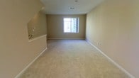 This two-story residence offers a straightforward and functional layout designed for comfortable everyday living. The interior is finished with wall-to-wall carpeting in a light, neutral tone that adds warmth underfoot and complements the beige walls found throughout the space. The living area is open and spacious, providing ample room for a variety of furniture arrangements. A centrally positioned window allows natural light to fill the room, creating a bright and airy atmosphere while offering a view of the surrounding greenery outside. The clean white baseboards and trim provide a crisp contrast to the warm wall tones, contributing to a cohesive and tidy aesthetic. Ceiling-mounted ventilation ensures consistent airflow throughout the interior. The two-floor configuration allows for a natural separation between living and private spaces, with the second level providing additional square footage and versatility. The overall design is understated and move-in ready, with neutral finishes that serve as a blank canvas for personalization. The space is well-suited for those seeking a practical, low-maintenance home with a calm and welcoming ambiance.