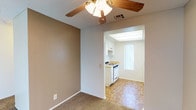 This single-floor residence offers a straightforward and functional layout designed for comfortable everyday living. The main living area features warm, neutral-toned walls and soft carpeted flooring, creating a welcoming atmosphere. A ceiling fan with an integrated light fixture provides both ventilation and ambient lighting, adding a practical touch to the space. The textured ceiling adds subtle character throughout. Adjacent to the living area, the kitchen is accessed through a wide archway, creating an open and connected feel between the two spaces. The kitchen is appointed with white cabinetry, a gas range, and a countertop workspace, all set atop classic parquet hardwood flooring that adds warmth and visual interest. A frosted vertical window in the kitchen allows natural light to filter in while maintaining privacy, and a recessed overhead light panel ensures the space remains bright and functional. White trim and baseboards run throughout, providing a clean contrast to the neutral wall tones. The overall ambiance is calm and practical, with a palette of soft beiges and whites that lend the space a timeless, move-in-ready quality.