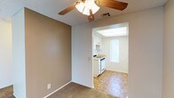 This single-floor residence offers a straightforward and functional layout designed for comfortable everyday living. The main living area features warm, neutral-toned walls and soft carpeted flooring, creating a welcoming atmosphere. A ceiling fan with an integrated light fixture provides both ventilation and ambient lighting, adding a practical touch to the space. The textured ceiling adds subtle character throughout. Adjacent to the living area, the kitchen is accessed through a wide archway, creating an open and connected feel between the two spaces. The kitchen is appointed with white cabinetry, a gas range, and a countertop workspace, all set atop classic parquet hardwood flooring that adds warmth and visual interest. A frosted vertical window in the kitchen allows natural light to filter in while maintaining privacy, and a recessed overhead light panel ensures the space remains bright and functional. White trim and baseboards run throughout, providing a clean contrast to the neutral wall tones. The overall ambiance is calm and practical, with a palette of soft beiges and whites that lend the space a timeless, move-in-ready quality.