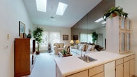 Located at 1110 E Cozza Dr in Spokane, Washington, this single-floor home offers a thoughtfully designed layout that balances comfort and functionality. The living room serves as the heart of the home, featuring soaring vaulted ceilings fitted with multiple skylights that flood the space with natural light. Large windows with vertical blinds frame views of the surrounding greenery, enhancing the bright and airy ambiance. An open-concept design connects the living area to the kitchen, which is equipped with light wood cabinetry, a white electric range, a dishwasher, and ample counter space. A peninsula with a sink further bridges the kitchen to the living space, making it ideal for everyday use. The dining area is distinguished by an elegant chandelier suspended from the vaulted ceiling, creating an inviting setting. A spacious bedroom includes sliding glass doors that open onto a private balcony with outdoor views, along with plush carpeting and generous natural light. The bathroom features a separate vanity area and a private water closet. Additional highlights include a large walk-in closet with wrap-around shelving and a flexible secondary room with direct access to the kitchen. Neutral tones and soft carpeting throughout lend the home a warm, cohesive feel.