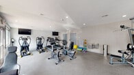 Located at 26 Cheswold Blvd in Newark, Delaware, this single-floor fitness center is thoughtfully designed to support a well-rounded workout routine. The space is finished with durable speckled flooring throughout, bright white walls, and recessed lighting that creates a clean, energizing atmosphere. Natural light filters in through large windows, offering views of the surrounding community.

The cardio section is well-appointed with multiple treadmills, elliptical trainers, and stationary bikes — including both upright and recumbent models — each accompanied by mounted flat-screen televisions for entertainment during workouts. A multi-station cable strength machine and an adjustable weight bench provide a solid foundation for resistance training. Supplementing the equipment is a Smart Fitness rack stocked with a variety of medicine balls and resistance accessories, along with foam rollers and exercise mats for stretching and functional training.

A built-in water fountain and stainless steel waste receptacles keep the space practical and tidy. Interior windows connect the gym to an adjacent room, adding a sense of openness. Multiple access points, including clearly marked exits, ensure easy entry and egress. Overall, the fitness center offers a comprehensive and accessible environment for residents to pursue their health and wellness goals.