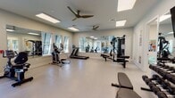 This fitness center is thoughtfully designed to support a comprehensive workout experience across a spacious, single-floor layout. The space is bathed in natural light through multiple windows fitted with adjustable blinds, while skylights and recessed panel lighting ensure consistent illumination throughout. Dual ceiling fans maintain comfortable airflow across the open exercise areas.

The main workout floor features light-toned tile flooring and is divided into distinct functional zones. The cardio area is equipped with treadmills, an elliptical trainer, and a stationary recumbent bike, all positioned to take advantage of the natural light and outdoor views through the windows. Large wall-to-wall mirrors amplify the sense of space and allow users to monitor their form during exercise.

A dedicated strength training section houses a multi-station cable machine, a weight bench, and a full rack of dumbbells ranging across various weights. A wall-mounted flat-screen television is positioned above the dumbbell rack for entertainment during workouts.

A separate flexibility and functional training area contains a rack stocked with stability balls in assorted sizes, medicine balls, and resistance mats. A recumbent bike and additional floor space make this zone ideal for stretching and low-impact exercise. Glass-paneled doors connect this area to adjacent community spaces, maintaining an open and connected atmosphere throughout the facility.