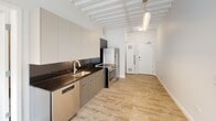Located in Chicago's Edgewater neighborhood, this single-floor unit at 1135 W Sheridan showcases a distinctly modern industrial aesthetic defined by its striking exposed beam ceiling. The white-painted structural beams run throughout the open layout, adding architectural character and a sense of generous vertical space.

The kitchen is thoughtfully designed with a galley-style configuration, featuring flat-front cabinetry in warm gray and cream tones, dark granite countertops with a matching backsplash, and a full suite of stainless steel appliances including a Beko refrigerator, electric range with oven, and dishwasher. Light hardwood-style flooring flows continuously throughout the unit, lending warmth and cohesion to each space.

The bedrooms are bright and minimalist, each equipped with large black-framed floor-to-ceiling windows that bring in natural light and frame views of the surrounding brick exterior. The exposed beam ceilings carry through these rooms, reinforcing the unit's consistent design language.

The bathroom features a clean, contemporary finish with large-format tile surrounds, a sliding glass-door walk-in shower, a light wood vanity, and a bold dark granite countertop. A dedicated closet space with wire shelving provides practical storage. Overall, the unit delivers a cohesive, modern aesthetic throughout.