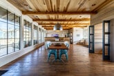 This single-floor leasing and community office for Vegina Apartment Villas presents a thoughtfully designed, open-plan interior that blends rustic and contemporary elements throughout. Visitors are welcomed through grand double wood-and-glass entry doors framed by large multi-pane windows that flood the entryway with natural light, complemented by warm hardwood flooring that extends throughout the entire space.

The main area features striking exposed wood beam ceilings that add architectural character and a sense of height overhead. A central communal dining table surrounded by vibrant teal metal chairs serves as a focal gathering point, anchored by an oversized industrial pendant light above. Adjacent to this area, a high-top bar counter with matching teal stools lines a feature wall accented with bold graphic signage, creating a casual collaborative zone.

Semi-private workstations are tucked behind teal-framed glass partition doors, offering a quieter workspace within the open environment. A compact, well-appointed kitchenette is finished with deep teal cabinetry, light stone countertops, a matte black faucet, and under-counter refrigeration — ideal for daily convenience. Additional seating nooks provide comfortable lounge areas throughout. The cohesive color palette of teal, natural wood, and white gives the space a modern yet inviting atmosphere.