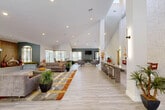 Located at 3495 Pinewalk Dr N in Margate, Florida, this single-story community clubhouse offers a thoughtfully designed, open-concept layout spanning a generous footprint. The interior is defined by soaring vaulted and double-height ceilings that create a bright, airy atmosphere, complemented by light-toned luxury vinyl plank flooring that flows seamlessly throughout the main areas.

The expansive main lounge serves as the centerpiece of the space, featuring plush sectional seating, a wood-paneled fireplace surround, and multiple seating arrangements suited for both social gatherings and relaxed lounging. Large windows along the perimeter flood the space with natural light and offer views of the surrounding landscaped grounds. Decorative potted plants and bold accent rugs add warmth and color to the contemporary design palette.

A long counter-height bar area with sleek pendant lighting provides additional seating, while a dedicated business center features multiple computer workstations, accent seating in vibrant yellow, and a stylish blue feature wall. Private office spaces with wrap-around windows and warm wood desks offer a quiet setting for one-on-one consultations. A beverage station with a wine refrigerator and a wall-mounted television complete the amenity offerings, making this clubhouse a fully equipped and welcoming community hub.