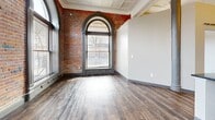 Located at 619 E William St in the heart of downtown Wichita, KS, this one-bedroom, one-bathroom unit blends historic industrial character with contemporary upgrades. The living area is an immediate standout, showcasing dramatic exposed brick walls, soaring ceilings with ornate coffered detailing, and large arched windows that flood the space with natural light and offer views of the surrounding streetscape. Rich, dark hardwood-style flooring runs throughout the main living areas, grounding the open layout with warmth.

The kitchen is well-appointed with stainless steel appliances — including a refrigerator, range, microwave, and dishwasher — complemented by dark granite countertops, white cabinetry, a textured tile backsplash, and open wood shelving. Exposed ductwork and recessed lighting reinforce the loft-inspired aesthetic.

A hallway with tall ceilings and bold dark-paneled doors leads to the bedroom and bathroom. The bedroom features carpeted flooring, a large ornately trimmed window with exposed brick surrounds, and a cozy, well-proportioned layout. The bathroom offers a full soaking tub-shower combination surrounded by earthy glazed stacked tile, wood-look flooring, and clean white fixtures, delivering a refined, spa-like feel within a compact footprint.