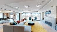 This thoughtfully designed single-floor space offers a seamless blend of modern aesthetics and functional amenities. The main lounge is an expansive, light-filled area anchored by a dramatic wall-to-wall floor-to-ceiling glass curtain system that frames sweeping outdoor views of the surrounding cityscape. A vibrant yellow area rug, a low-profile round coffee table, and a generously sized sectional sofa with colorful accent pillows create a welcoming focal point. Flanking the room is a striking feature wall housing a linear gas fireplace set beneath a marble-clad surround and a large wall-mounted television. Built-in bench seating with upholstered cushions and decorative paneling lines both sides of the fireplace, adding both comfort and architectural detail. Curated artwork and open shelving with dark wood cabinetry add warmth and character throughout. Adjacent to the lounge, a dedicated game room features light hardwood flooring, a full-size ping-pong table, and ample natural light from large windows overlooking the outdoors. Warm cove lighting along the tray ceiling adds an inviting ambiance. The overall aesthetic is contemporary and polished, with a consistent palette of white, grey, and wood tones accented by bold pops of teal and gold.