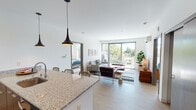 This single-floor residence showcases a thoughtfully designed open-concept layout that seamlessly connects the kitchen, dining, and living areas. The kitchen is anchored by a generous granite-topped island with bar seating for three, complemented by white cabinetry, stainless steel appliances, and sleek pendant lighting overhead. Light wood-tone flooring flows consistently throughout the space, enhancing the unit's cohesive, modern aesthetic.

The living area is bright and welcoming, with large sliding glass doors that open onto a private balcony offering outdoor views of surrounding greenery. Natural light pours in generously, contributing to the airy atmosphere of the main living space.

Two well-proportioned bedrooms provide comfortable retreats. One bedroom features floor-to-ceiling windows with expansive neighborhood views, while the other offers a quieter setting with access to its own space. Both rooms feature the same light wood flooring found throughout.

The two bathrooms are finished with granite countertops, floating white vanities, large mirrors, and combination tub-shower units. A hallway with wood-paneled doors connects the private areas of the unit, completing a clean, contemporary floor plan designed for comfortable, functional living.
