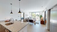 This single-floor residence showcases a thoughtfully designed open-concept layout that seamlessly connects the kitchen, dining, and living areas. The kitchen is anchored by a generous granite-topped island with bar seating for three, complemented by white cabinetry, stainless steel appliances, and sleek pendant lighting overhead. Light wood-tone flooring flows consistently throughout the space, enhancing the unit's cohesive, modern aesthetic.

The living area is bright and welcoming, with large sliding glass doors that open onto a private balcony offering outdoor views of surrounding greenery. Natural light pours in generously, contributing to the airy atmosphere of the main living space.

Two well-proportioned bedrooms provide comfortable retreats. One bedroom features floor-to-ceiling windows with expansive neighborhood views, while the other offers a quieter setting with access to its own space. Both rooms feature the same light wood flooring found throughout.

The two bathrooms are finished with granite countertops, floating white vanities, large mirrors, and combination tub-shower units. A hallway with wood-paneled doors connects the private areas of the unit, completing a clean, contemporary floor plan designed for comfortable, functional living.