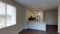 This single-floor space showcases a thoughtfully designed open-concept layout that seamlessly connects the kitchen and living areas. The kitchen is a standout feature, anchored by a wide white peninsula with a light-colored countertop that provides ample prep space and doubles as a casual dining or gathering area. White upper cabinetry lines the kitchen walls, offering generous storage while maintaining a clean, bright aesthetic. Stainless steel appliances, including a full-sized refrigerator, contribute a modern, professional feel, and a dark tile backsplash adds a striking contrast against the white surfaces. Two sleek pendant lights hang above the kitchen area, providing focused task lighting and an elegant design touch. Recessed ceiling lights further illuminate the space. Dark hardwood-style flooring runs throughout, grounding the room with warmth and depth while complementing the neutral white walls. A window fitted with white horizontal blinds on the adjacent wall invites natural light into the living area, enhancing the overall brightness of the space. The combination of contemporary finishes and an open, airy layout creates a functional and visually cohesive environment.