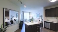 This single-floor residence offers a well-designed open-concept layout that seamlessly connects the kitchen and living areas. The kitchen is a standout feature, anchored by a large island with rich dark cabinetry, sleek chrome hardware, and a crisp white countertop that provides ample prep and bar-style seating space. Track lighting overhead ensures the kitchen is well-lit and functional, complementing the natural light that pours in from the generously sized windows throughout the space. The living area flows naturally from the kitchen, creating an inviting and cohesive entertaining space. Large windows dressed with elegant drape panels flood the room with abundant natural light, enhancing the airy, open ambiance. The hard-surface flooring throughout adds a contemporary touch and contributes to the cohesive aesthetic of the home. Lush greenery and thoughtfully placed decor bring warmth and life to the interior. A large decorative mirror near the entryway amplifies the sense of space. Overall, this residence balances modern design with a warm, livable atmosphere, offering a comfortable and stylish environment throughout its single level.