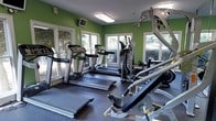 This fitness center is a single-floor facility designed to support a comprehensive workout experience. The space is outfitted with a range of strength training equipment, including a multi-station cable and weight machine ideal for a variety of resistance exercises. Cardio options are also well represented, with a rowing machine and a stationary bike positioned near the windows, allowing users to exercise with a view of the surrounding exterior. The flooring throughout is durable rubber matting, providing cushioning and stability during workouts. Overhead fluorescent lighting ensures the space is well-lit, complemented by natural light streaming in through multiple windows along the walls, creating a bright and energizing atmosphere. A wall-mounted television is positioned to keep users entertained during their sessions. The clean, white walls contribute to an open and airy feel. An entry door and informational signage are also present, adding to the room's functional layout. Overall, the fitness center offers a practical and accessible environment suited for both strength training and cardiovascular exercise.