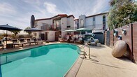 Located at 4341 Spring St in La Mesa, California, this outdoor amenity space showcases a resort-style environment designed for relaxation and recreation. The centerpiece is a generously sized, kidney-shaped swimming pool with clear turquoise water, framed by a spacious sun deck finished in light-toned concrete pavers. Multiple lounge chairs and sun umbrellas are arranged throughout the deck, providing ample space for sunbathing and outdoor leisure.

Adjacent to the main pool, a separate circular hot tub offers a dedicated space for hydrotherapy and relaxation. The surrounding patio area is further enhanced by seating arrangements with tables and chairs shaded by umbrellas, ideal for casual outdoor gathering.

The space is enclosed by a multi-story building featuring Spanish Colonial Revival architecture, characterized by stucco exteriors, clay tile rooflines, arched detailing, and wrought iron balcony railings. Private balconies overlook the pool courtyard, adding an elevated sense of community to the outdoor setting. A decorative wood-paneled privacy fence lines one boundary of the pool area, adorned with circular wall art and backed by mature eucalyptus trees that provide natural shade and a lush green backdrop. The surrounding hillside views and the abundance of natural Southern California sunlight complete the inviting, open-air atmosphere of this well-appointed outdoor space.