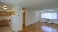This single-floor residence offers a straightforward and functional layout designed for comfortable everyday living. The living area is finished with warm hardwood flooring that extends throughout the space, contributing to a cohesive and inviting atmosphere. Bright white walls keep the interior feeling clean and open, while a window along the far wall allows natural light to fill the room, enhancing the sense of space. A baseboard heating unit is positioned beneath the window, providing efficient climate control. Adjacent to the living area, the kitchen is partially visible and features light wood cabinetry and a white refrigerator, offering a practical and accessible cooking space. A closet is conveniently located between the living and kitchen areas, providing built-in storage. Smoke detectors are installed on the ceiling, and multiple electrical outlets and switches are thoughtfully placed throughout. The overall ambiance is understated and neutral, making the space a versatile canvas well-suited to a variety of furnishing styles and personal tastes.