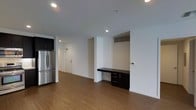 This single-floor space showcases a clean, contemporary design with an open-concept layout that seamlessly connects the kitchen and living areas. The kitchen is equipped with stainless steel refrigerator and dark cabinetry, offering both style and functionality. Warm hardwood-style flooring extends throughout the unit, adding a welcoming feel to the bright, white interior.

A standout feature is the built-in desk nook, complete with dark wood cabinetry and drawers, making it an ideal dedicated workspace integrated directly into the living area. Above the nook, a ventilation grille is neatly recessed into the wall, maintaining the clean lines of the design. Recessed lighting in the ceiling provides consistent, even illumination across the space.

The layout offers a clear sightline from the main living area down the hallway toward the entryway, creating a sense of depth and openness. White walls and crisp baseboards throughout contribute to a minimalist, modern aesthetic. The entry door features a standard door frame with adjacent light switches, keeping the design functional and uncluttered. Overall, the space presents a practical yet stylish environment with thoughtful built-in details and a cohesive modern finish palette.