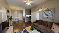 This single-story unit is designed with a functional, open layout that promotes a comfortable and connected living experience. The living room serves as the central hub of the space, featuring vaulted ceilings that lend an airy, expansive feel. Rich hardwood-style flooring runs throughout the main living areas, complemented by neutral gray walls that create a clean, modern aesthetic. A ceiling fan provides additional comfort, while a large sliding glass door floods the room with natural light and provides access to the outdoors.

The kitchen is efficiently laid out and equipped with white cabinetry, white appliances including a range, microwave, dishwasher, and refrigerator, along with light countertops. A pass-through counter connects the kitchen to the adjacent dining area, enhancing the open flow of the space.

The bedroom offers a comfortable retreat with plush carpeting, a window with blinds for natural light control, and ample wall space. The full bathroom includes a single-sink vanity with gray cabinetry, a large mirror, and a shower/tub combination. A dedicated laundry area with wire shelving and washer hookups rounds out the unit's practical amenities.