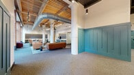 This expansive single-floor amenity space blends industrial character with modern design, offering a thoughtfully curated environment across multiple functional zones. The open layout is anchored by soaring ceilings that expose warm wood decking, structural beams, and large-diameter ductwork — a nod to the building's industrial heritage — all illuminated by a combination of track lighting, pendant fixtures, and bold oval ring chandeliers.

The main lounge area features multiple seating arrangements with leather sofas, accent chairs, and stylish side tables set atop patterned area rugs, creating inviting spaces for socializing or relaxing. Floor-to-ceiling black-framed windows along the street-facing wall flood the interior with natural light and offer unobstructed urban views, framed by exposed brick. Teal board-and-batten wainscoting adds a striking design detail near the entry, complementing the space's eclectic palette.

A sleek wet bar with dark vertical tile backsplash, sage cabinetry, and a long white countertop with bar seating anchors one side of the space, alongside a bank of package lockers. Glass-walled partitions lead to a dedicated fitness center with cardio equipment visible from the corridor. Additional rooms include a flexible space with light wood flooring and warm-toned walls. A connecting hallway ties all areas together seamlessly.