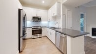 This single-floor residence offers a clean, modern aesthetic defined by light-toned hardwood flooring, neutral gray walls, and white trim throughout. The open-concept living area flows seamlessly into a compact dining space, highlighted by crown molding, recessed lighting, and a gently curved architectural accent wall that adds character to the layout. A sliding glass door in one area invites natural light and provides views of the surrounding neighborhood.

The kitchen is well-appointed with white shaker cabinetry, dark quartz countertops, a subway tile backsplash, and a full suite of stainless steel appliances including a gas range, built-in microwave, dishwasher, and French door refrigerator. A peninsula counter offers additional prep and seating space.

The bedrooms are generously sized, each featuring wide-plank hardwood flooring and large windows that frame lush green tree canopies, filling the rooms with natural light. One bedroom includes a French door leading to an exterior area.

Two bathrooms mirror each other in design, each featuring white shaker vanity cabinets, dark quartz counters, undermount sinks, and LED backlit vanity mirrors. Both include a full tub-shower combination with clean white surrounds. The overall ambiance is bright, fresh, and move-in ready.