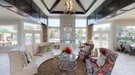 This single-story clubhouse is designed with an open, airy layout that blends contemporary elegance with resort-style comfort. The centerpiece of the space is a dramatic vaulted ceiling with bold dark beam detailing and a geometric chandelier, creating a striking architectural focal point. Expansive floor-to-ceiling windows and multiple sets of French doors wrap the main lounge area, flooding the interior with natural light and offering unobstructed views of the outdoor swimming pool and palm-lined surroundings.

The main lounge is furnished with a curved ivory sofa, vibrant patterned accent chairs, and a geometric glass coffee table atop a plush area rug. A built-in water feature with the community logo serves as a decorative backdrop. Stone accent columns add texture and warmth throughout.

A dedicated café-style kitchenette features dark cabinetry, light countertops, a wall-mounted TV, a reflective mosaic tile backsplash, and a compact refrigerator for resident convenience. The entryway corridor features travertine tile flooring, sheer drapery panels, and a tufted bench seat. Interior transitions are handled through a modern frosted-glass sliding barn door, leading to restroom facilities. Dining seating near the poolside doors completes the space.